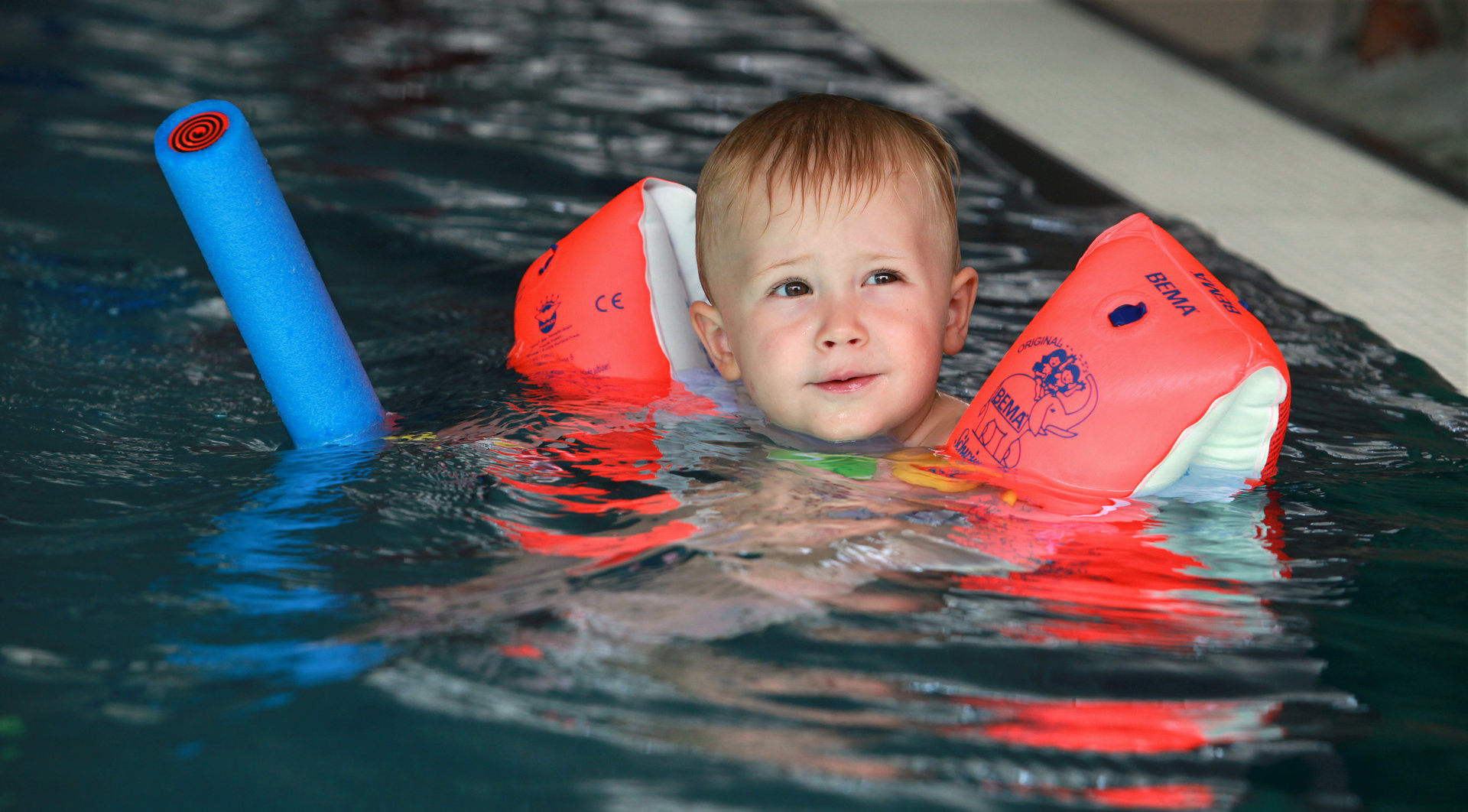 ein kleiner  blonder Junge mit Schwimmflügeln und einer Schwimmnudel planscht im Kleinkinderbecken
