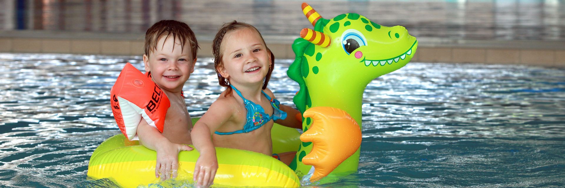 ein kleiner Junge mit Schwimmflügeln und ein kleines Mädchem in türkisenen Bikini sitzen auf einem grünen Luftballontier im Kleinkinderbecken