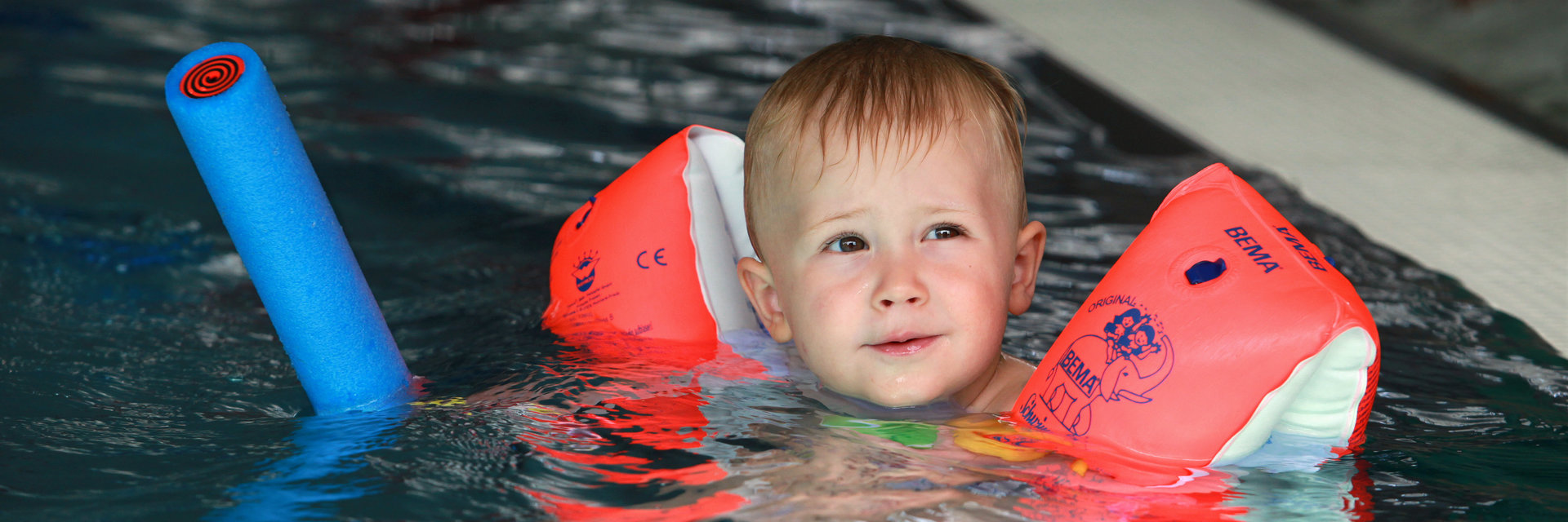 ein kleiner  blonder Junge mit Schwimmflügeln und einer Schwimmnudel planscht im Kleinkinderbecken